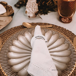 Um guardanapo branco, com anel personalizado "MARGARIDA", repousa sobre um prato cinzento, em cima de um individual rústico. Ao fundo, detalhes de decoração de casamento e talher dourado. Um guardanapo branco, com anel personalizado "MARGARIDA", repousa sobre um prato cinzento, em cima de um individual rústico. Ao fundo, detalhes de decoração de casamento e talher dourado.
