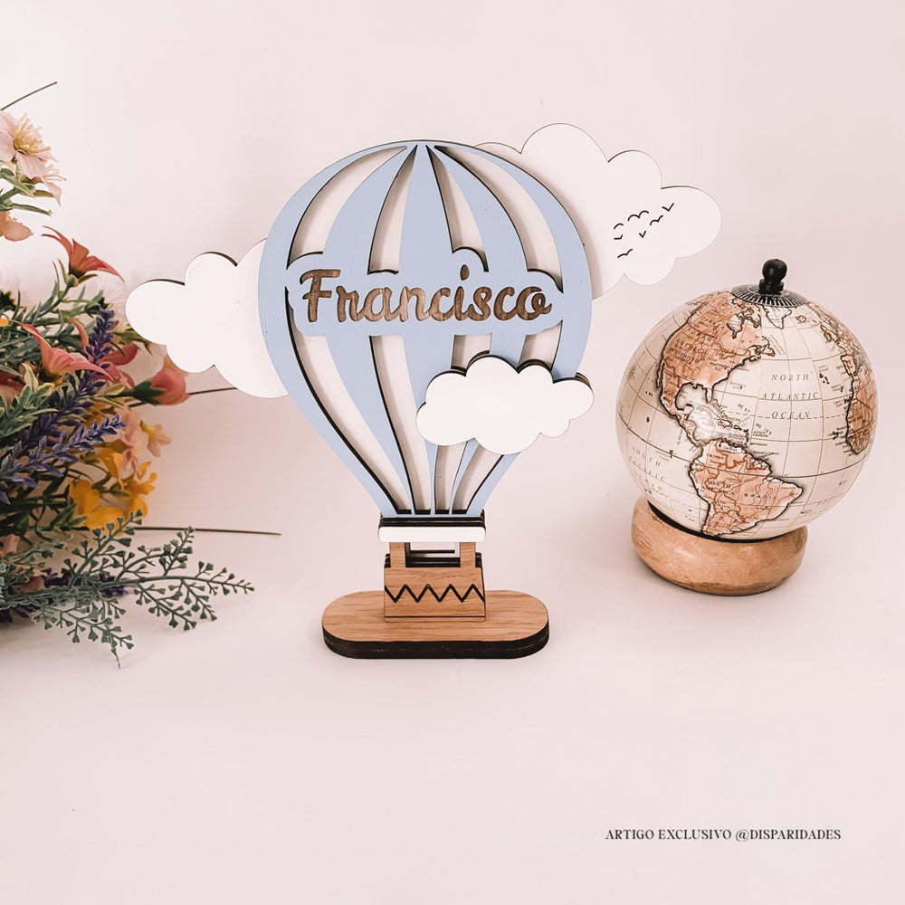 Balão de ar quente decorativo com nome "Francisco" e nuvens, ao lado de um globo terrestre e flores, estilo rústico.
