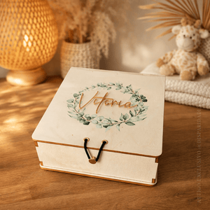 Caixa de madeira com nome “Vitória” em relevo dourado e coroa de folhas sobre mesa, com luz suave Caixa de madeira com nome “Vitória” em relevo dourado e coroa de folhas sobre mesa, com luz suave