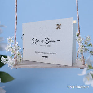 Capa para livro de honra em branco personalizado como o nome dos noivos, data e mensagem, tema viagens, colocada sobre um baloiço com flores brancas e um fundo azul Capa para livro de honra em branco personalizado como o nome dos noivos, data e mensagem, tema viagens, colocada sobre um baloiço com flores brancas e um fundo azul