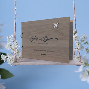 Capa para livro de honra em carvalho personalizado como o nome dos noivos, data e mensagem, tema viagens, colocada sobre um baloiço com flores brancas e um fundo azul Capa para livro de honra em carvalho personalizado como o nome dos noivos, data e mensagem, tema viagens, colocada sobre um baloiço com flores brancas e um fundo azul
