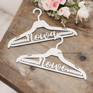 Cabides brancos de madeira com a palavra "Noiva" escrita em letras cursivas. Estão sobre uma mesa de madeira rústica, ao lado de um pequeno ramo de flores cor-de-rosa e brancas. Cabides brancos de madeira com a palavra "Noiva" escrita em letras cursivas. Estão sobre uma mesa de madeira rústica, ao lado de um pequeno ramo de flores cor-de-rosa e brancas.