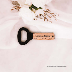 Abre-caricas de madeira com "Cátia & Bruno" e data, sobre tecido branco com flores e ramos secos, criando um cenário rústico e elegante. Abre-caricas de madeira com "Cátia & Bruno" e data, sobre tecido branco com flores e ramos secos, criando um cenário rústico e elegante.