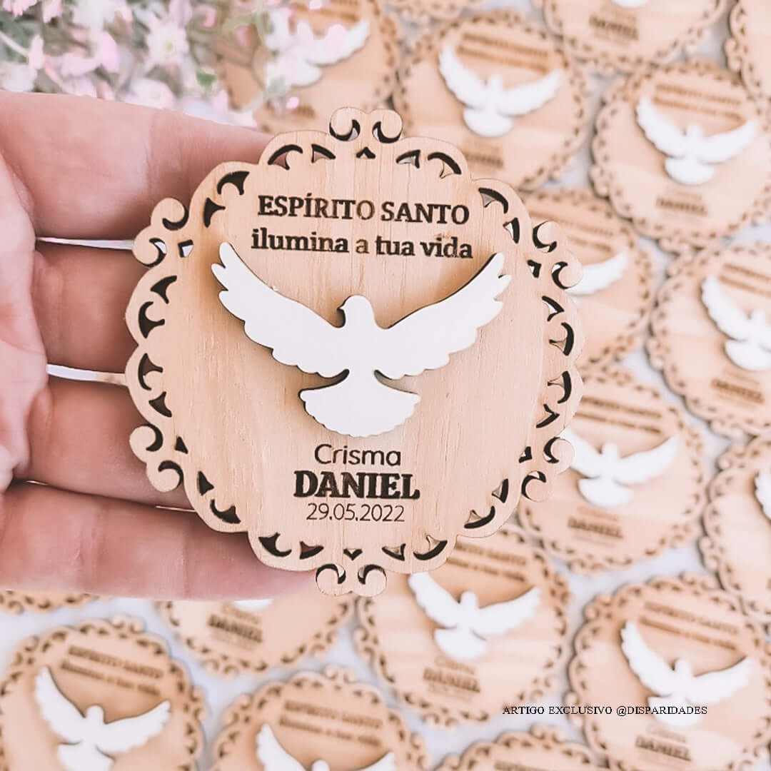 A mão segura uma lembrança de madeira em forma de arabesco com detalhes vazados e um desenho de pomba no centro, simbolizando o Espírito Santo, com a inscrição "ESPÍRITO SANTO ilumina a tua vida Crisma DANIEL 29.05.2022". Ao fundo, vários recortes similares estão desfocados A mão segura uma lembrança de madeira em forma de arabesco com detalhes vazados e um desenho de pomba no centro, simbolizando o Espírito Santo, com a inscrição "ESPÍRITO SANTO ilumina a tua vida Crisma DANIEL 29.05.2022". Ao fundo, vários recortes similares estão desfocados