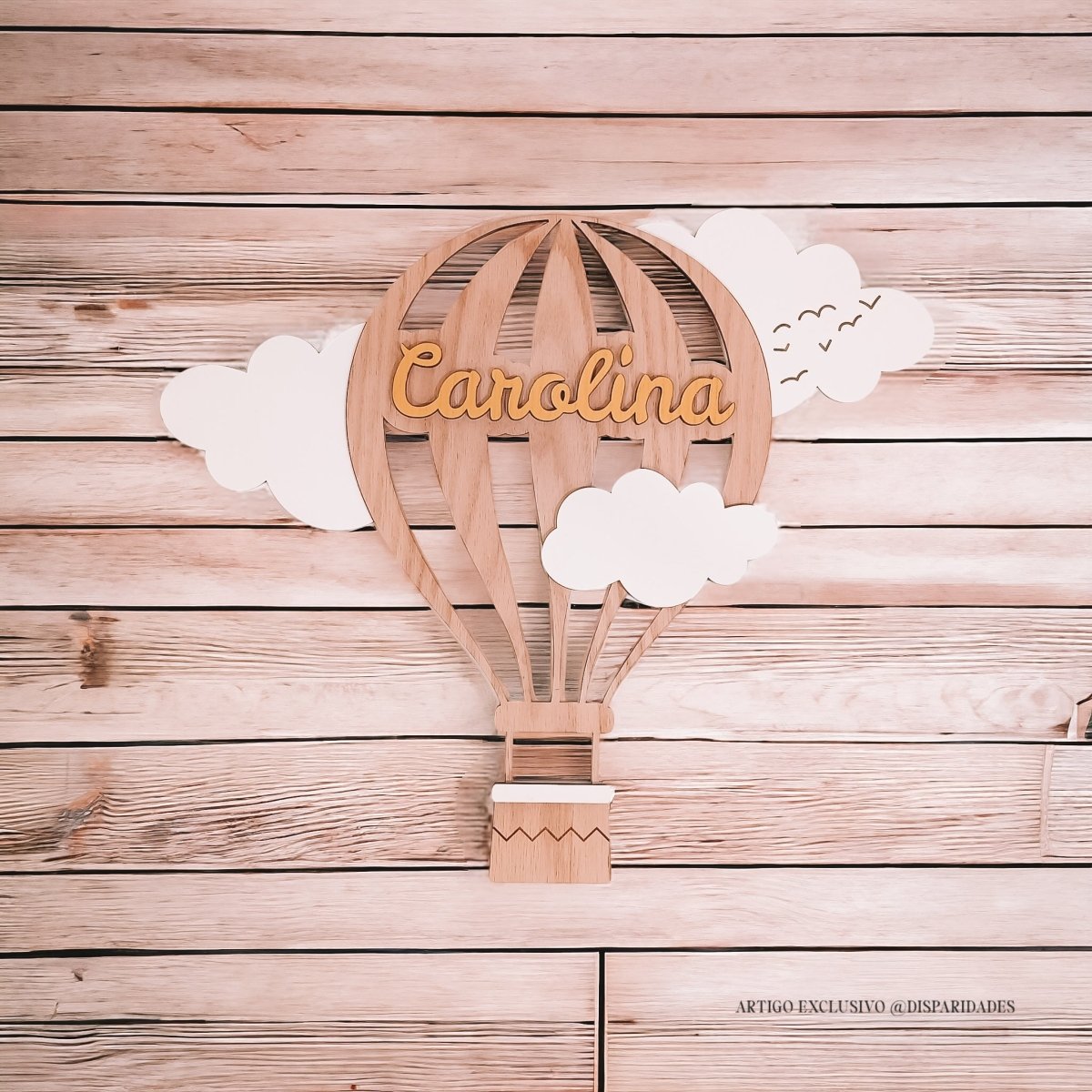 Decoração de parede rústica com um balão de ar quente em madeira, onde se lê “Carolina”, cercado por nuvens brancas, contra um fundo de tábuas de madeira claras.