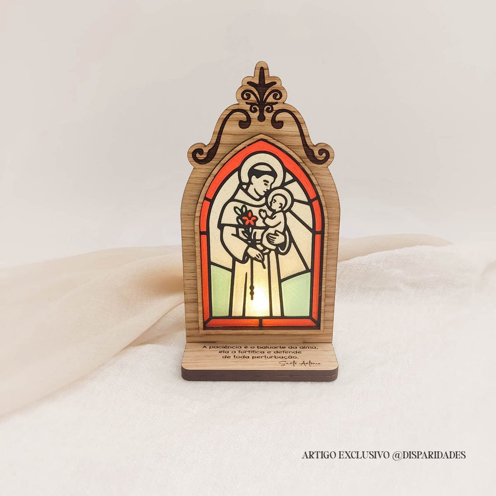 Oratório rústico com vitral iluminado de Santo António a segurar o Menino Jesus, com frase sobre paciência gravada na base em madeira.