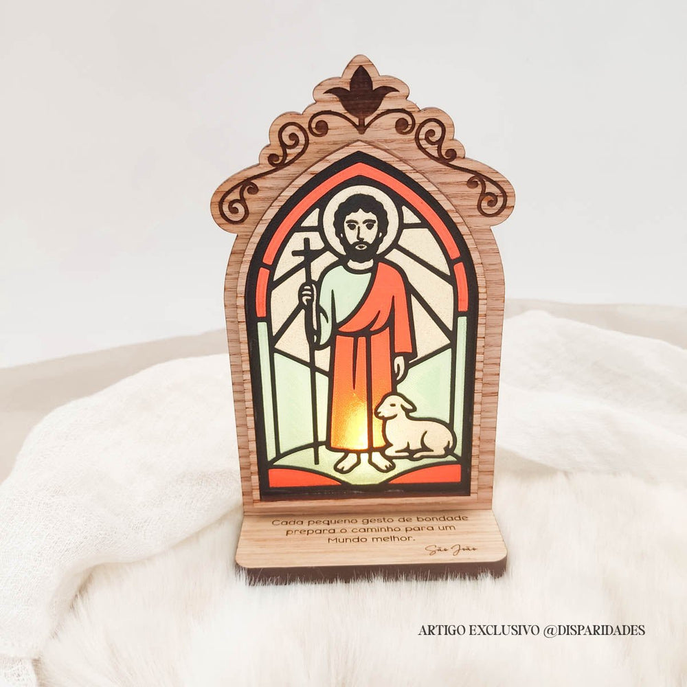 Oratório rústico iluminado com imagem de São João em vitral, com ovelha aos pés e frase: “Cada pequeno gesto de bondade prepara o caminho para um mundo melhor.