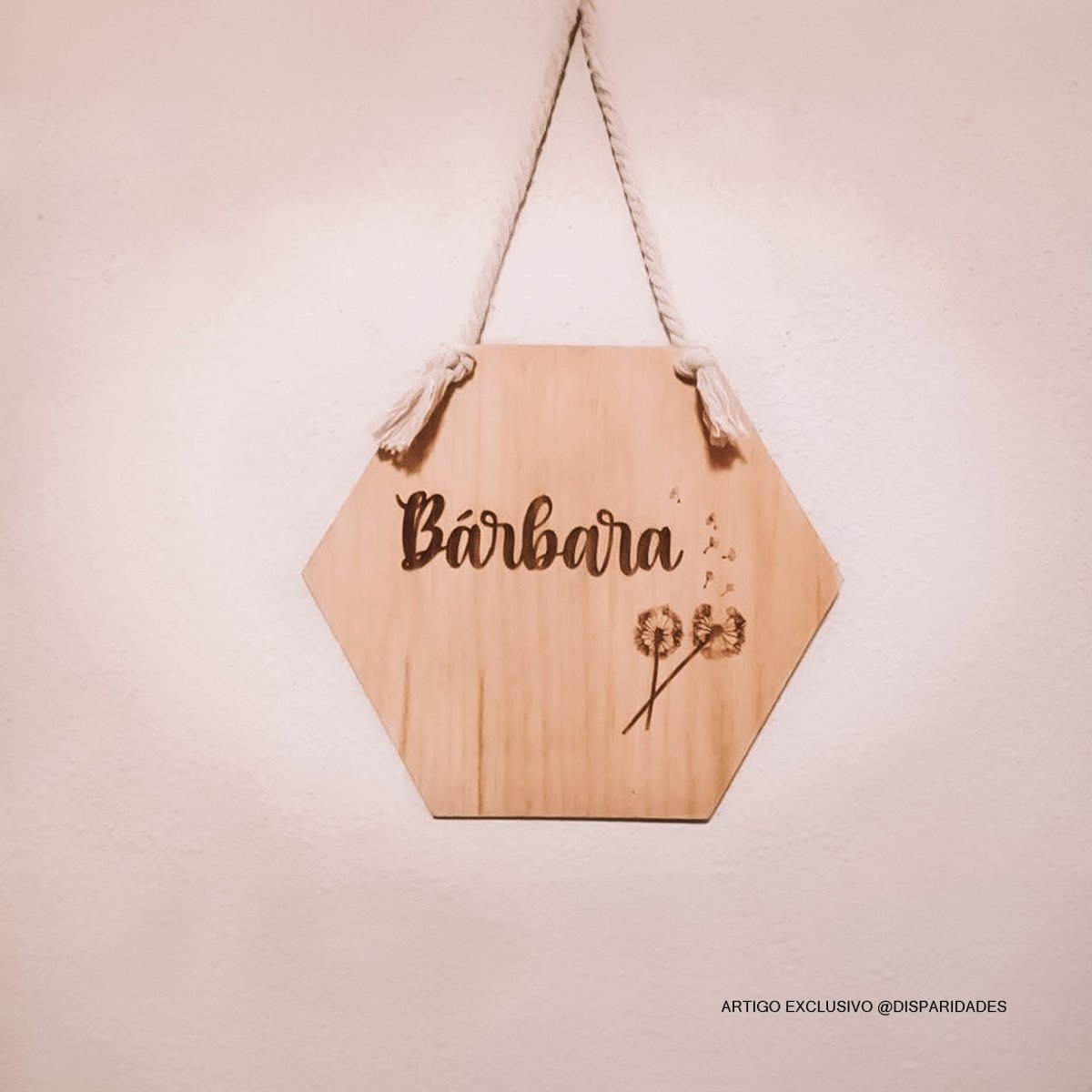 Placa decorativa de madeira natural com forma hexagonal, pendurada por uma corda com nós nas extremidades. O nome 'Bárbara' está gravado centralmente em letras cursivas com destaque. No canto inferior direito, um desenho delicado de um dente-de-leão é também gravado na madeira.