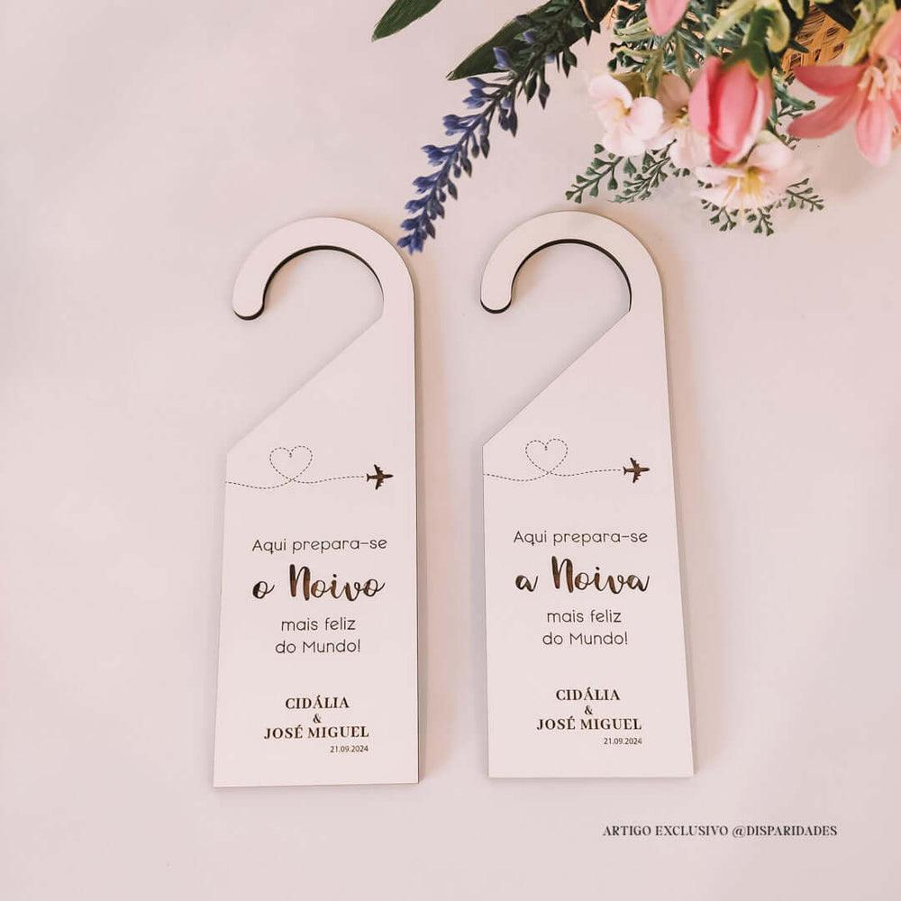 Placas de cabide brancas com texto para casamento. À esquerda, lê-se "Aqui prepara-se o Noivo mais feliz do Mundo!" e à direita "Aqui prepara-se a Noiva mais feliz do Mundo!", com nomes Cidália e José Miguel e a data 21.09.2024. Decoradas com flores e ramos.