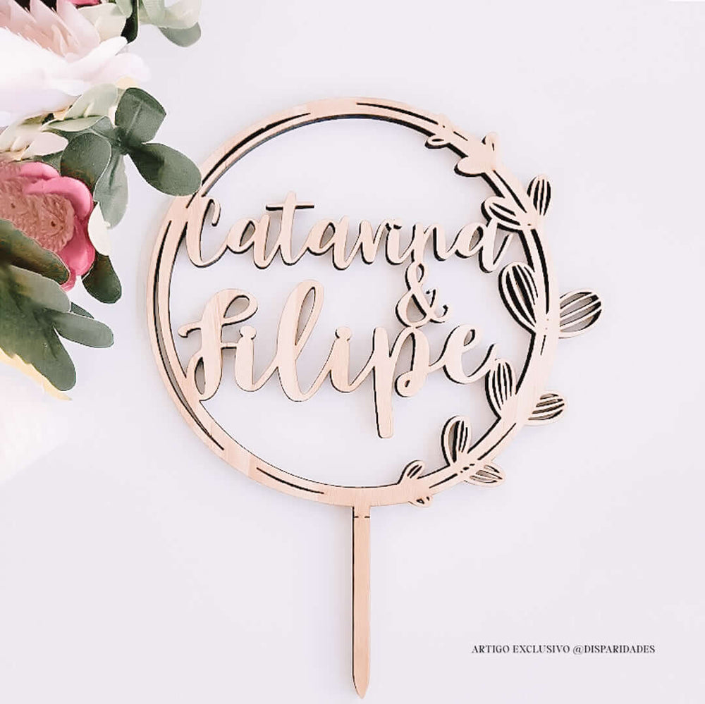 Topo de bolo em madeira rústica personalizado com os nomes "Catarina & Filipe" dentro de um círculo decorado, acompanhado de flores recortadas