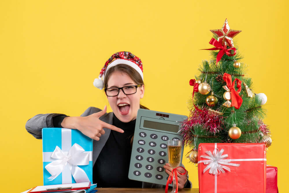 uma mulher com uma calculadora em uma mão e a outra a apontar, ao seu redor presentes e uma árvore de natal
