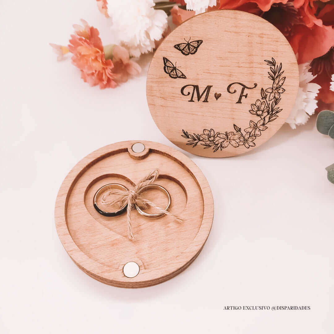Porta-alianças aberto em madeira rústica com alianças amarradas, tampa gravada com iniciais "M♥F", borboletas e flores, sobre fundo floral.