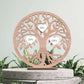 Árvore da vida em madeira com recortes de folhas e corações contendo os nomes Alice, Diana, Camila, sobre pedestal de concreto, com folhagem ao fundo.