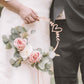 Casal segurando decoração de casamento 'you & me' em madeira com flores e folhas de eucalipto.