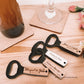  Abre-caricas de madeira personalizados com nomes e datas, sobre uma mesa com detalhes rústicos, junto a uma garrafa e copo com base de madeira.