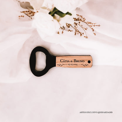 Abre-caricas de madeira com "Cátia & Bruno" e data, sobre tecido branco com flores e ramos secos, criando um cenário rústico e elegante.