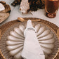 Um guardanapo branco, com anel personalizado "MARGARIDA", repousa sobre um prato cinzento, em cima de um individual rústico. Ao fundo, detalhes de decoração de casamento e talher dourado.