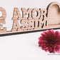 Moldura rústica com frase "O AMOR É ASSIM" em madeira, ao lado de uma flor vermelha, sobre fundo branco.