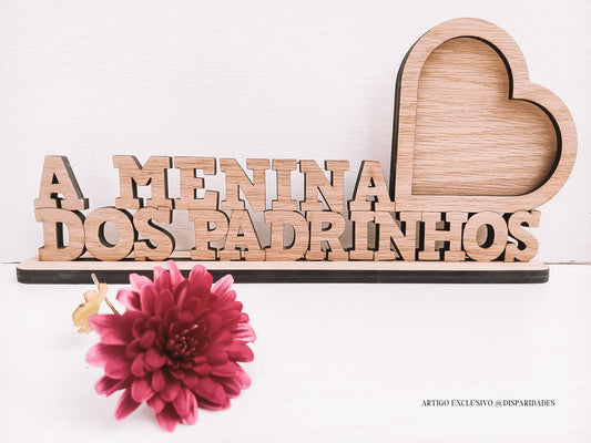 Letras em madeira formam "A MENINA DOS PADRINHOS" com um coração e uma flor rosa em primeiro plano.