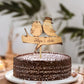 Topo de bolo com dois pássaros estilizados em madeira, usando chapéu e flor, segurando faixa "Filipa & João". Bolo de chocolate polvilhado com açúcar em prato branco.