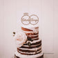 Bolo de casamento com três andares, cobertura branca e camadas visíveis de bolo castanho, decorado com uma rosa pálida e folhas verdes. No topo, destaca-se um Topo de bolo com os nomes "Vera & Tiago" e duas alianças entrelaçados.