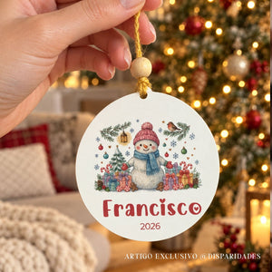 Mão a segurar uma bola de Natal personalizada com boneco de neve, nome Francisco e ano 2026, com árvore iluminada ao fundo.

