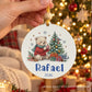 Mão a segurar uma bola de Natal com urso, pinheiro e presentes, nome Rafael e ano 2026, com árvore iluminada ao fundo.
