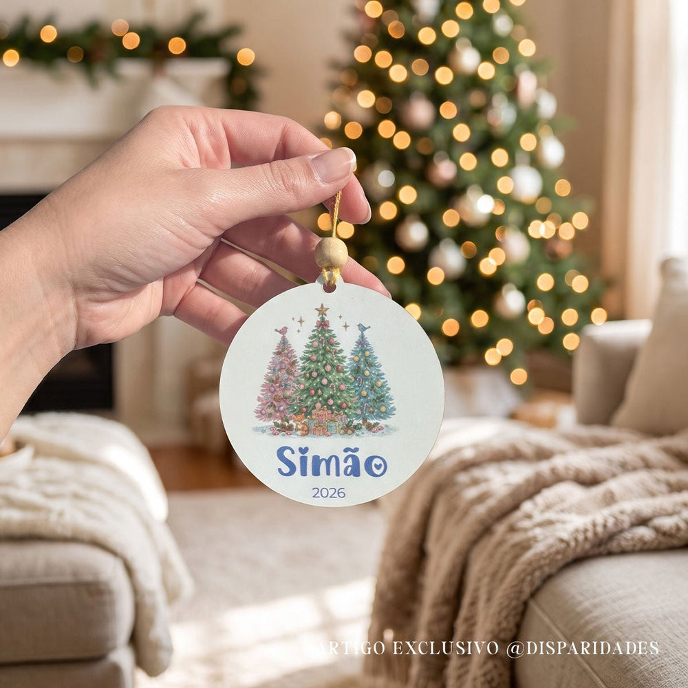 Mão a segurar uma bola de Natal com três pinheiros decorados, nome Simão e ano 2026, com árvore iluminada ao fundo.
