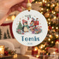 Mão a segurar bola de Natal com pinguim, caixa de correio e presentes, nome Tomás e ano 2026, com árvore iluminada ao fundo.

