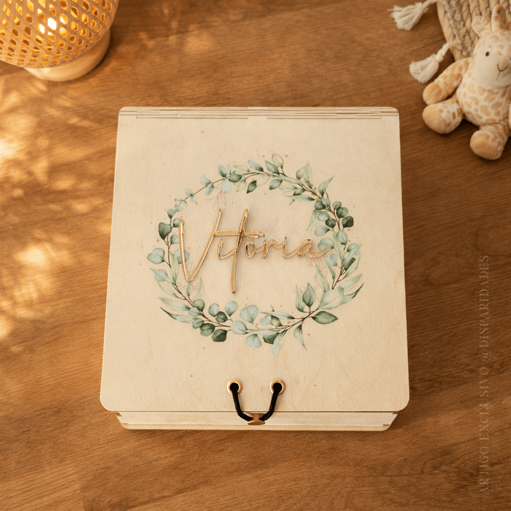 Caixa de madeira personalizada com nome “Vitória” em dourado e coroa de folhas, sobre mesa com iluminação suave
