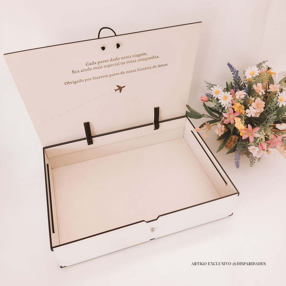 Caixa aberta de cor clara com texto sentimental e avião desenhado, ao lado de um bouquet rústico de flores silvestres. Estilo minimalista e elegante.