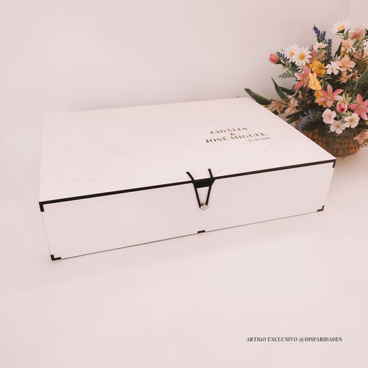 Caixa fechada branca com fecho preto e nomes personalizados, acompanhada de um arranjo rústico de flores variadas. Estilo simples e personalizado.