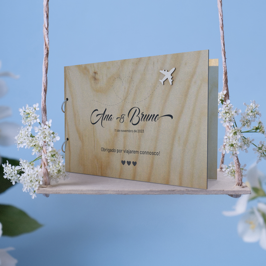 Capa para livro de honra em branco personalizado como o nome dos noivos, data e mensagem, tema viagens, colocada sobre um baloiço  com flores brancas e um fundo azul