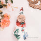 argola para guardanapos rústico em madeira com iniciais e desenho de flor, sobre guardanapo floral colorido, com flores artificiais ao redor.