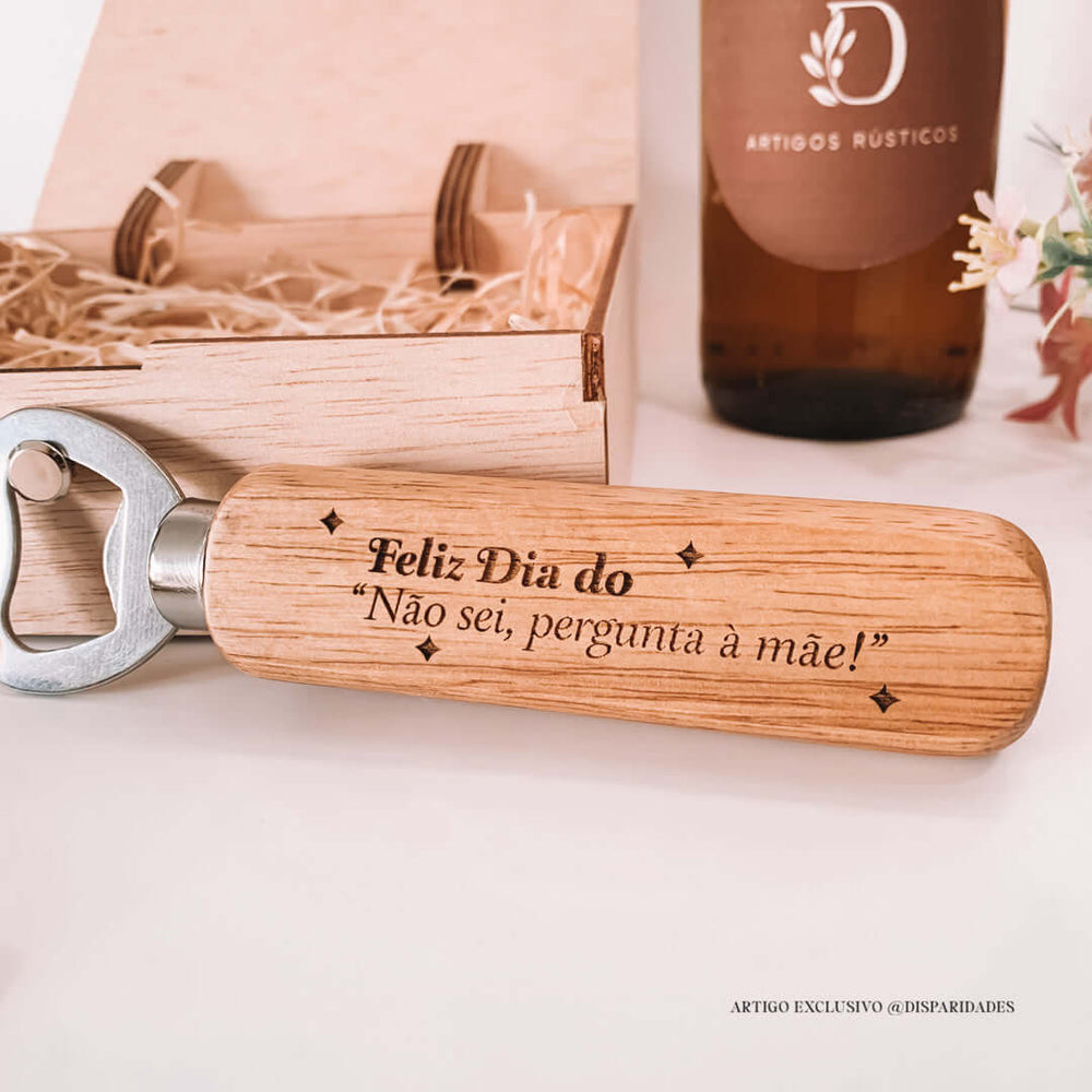 Um abre-caricas rústico em madeira com gravação "Feliz Dia do 'Não sei, pergunta à mãe!'", ao lado de uma caixa de madeira e uma garrafa com etiqueta de artigos rústicos.