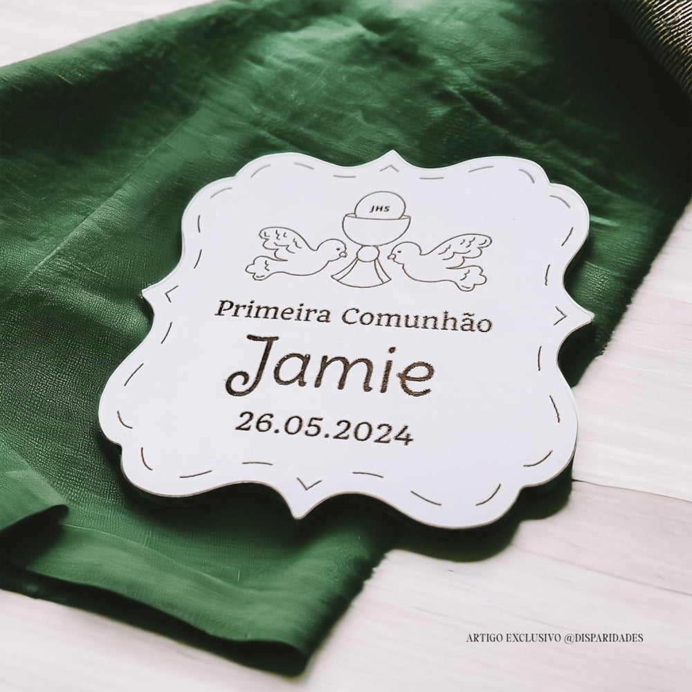 Uma lembrança personalizada da Primeira Comunhão, com nome e a data da cerimónia gravados numa placa de madeira branca. A placa está decorada com um pano verde com um padrão floral e a inscrição "JHS".