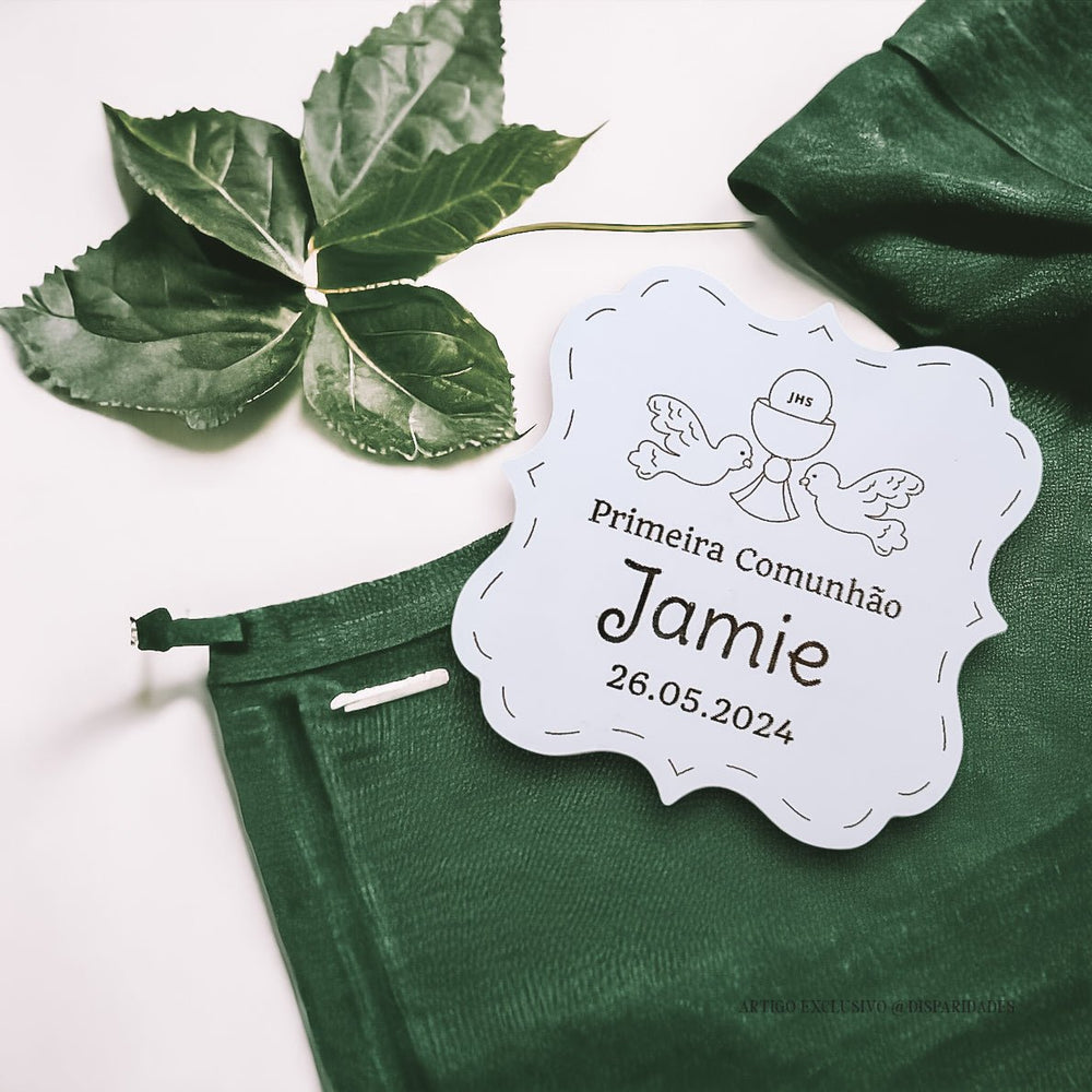 Uma lembrança personalizada da Primeira Comunhão, com nome e a data da cerimónia gravados numa placa de madeira branca. A placa está decorada com um pano verde com um padrão floral e a inscrição "JHS"