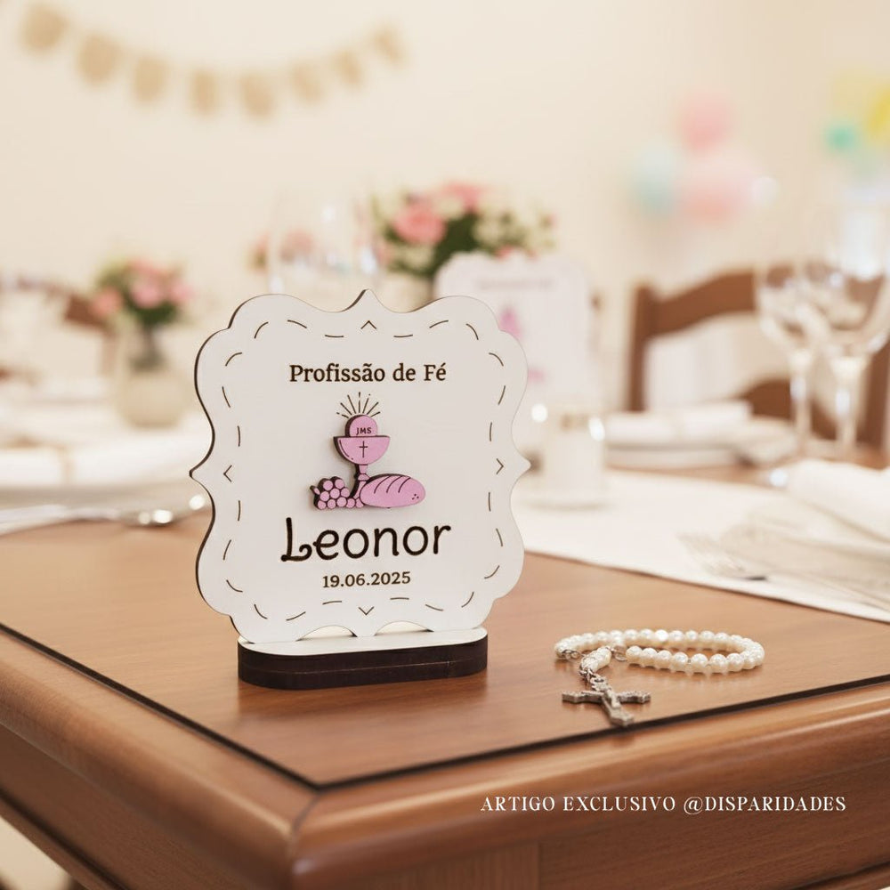 lembrança com base gravado Profissão de fé com o nome Leonor e data. A lembrança está em cima de uma mesa ao lado de um terço