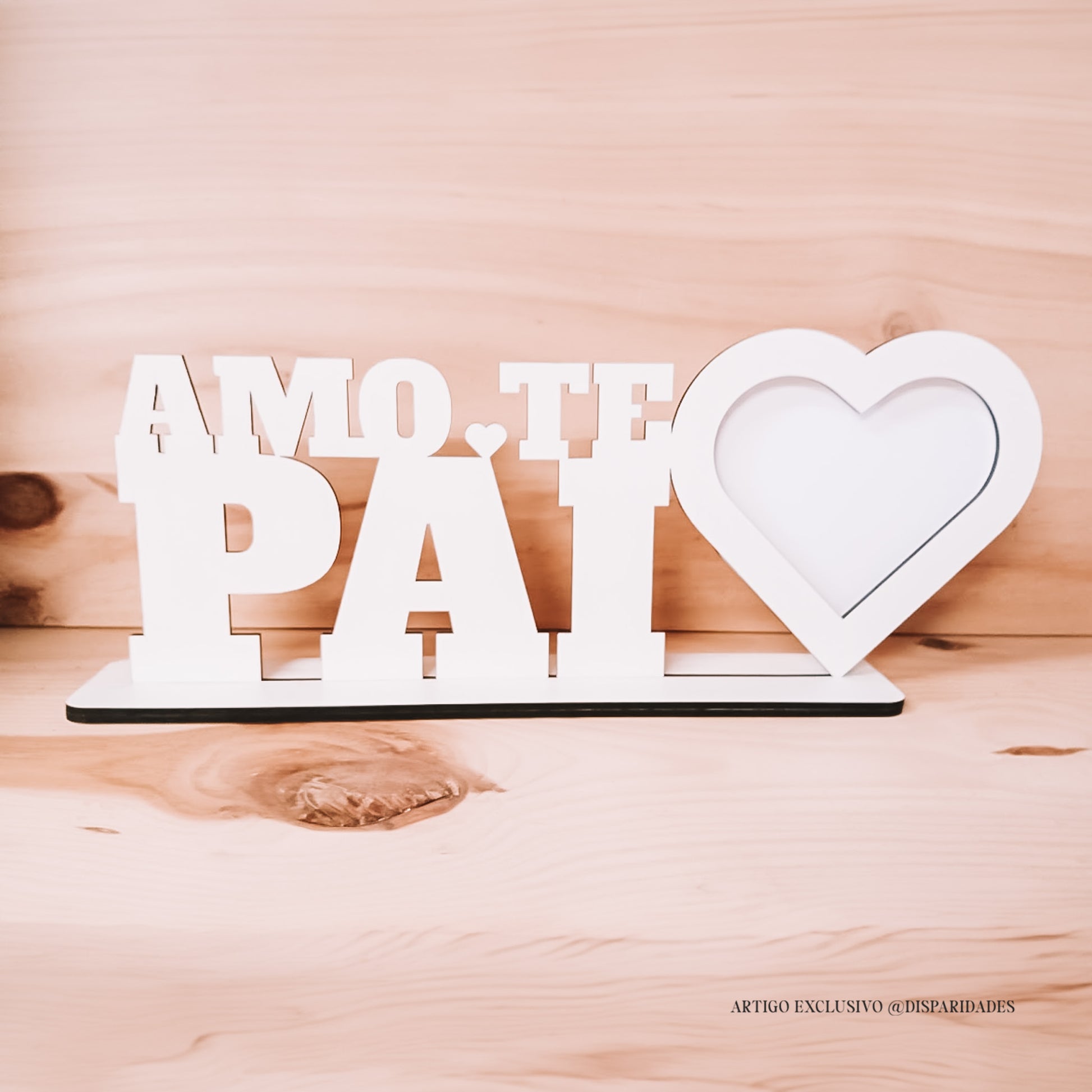 Moldura em branco com "AMO-TE PAI" e coração, sobre base branca, contrastando com fundo de madeira claro.