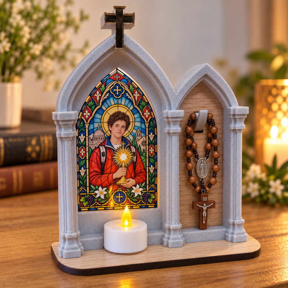 Oratório de São Carlo Acutis com vitral colorido e vela LED acesa, com terço de madeira pendurado no suporte sobre a dedicatória gravada.
