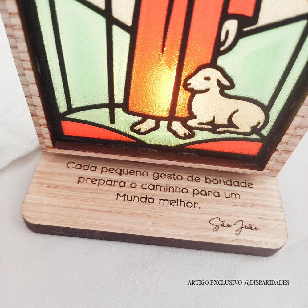 Oratório rústico iluminado com imagem de São João em vitral, com ovelha aos pés e frase: “Cada pequeno gesto de bondade prepara o caminho para um mundo melhor.