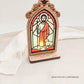 Oratório rústico iluminado com imagem de São João em vitral, com ovelha aos pés e frase: “Cada pequeno gesto de bondade prepara o caminho para um mundo melhor.