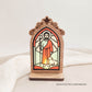 Oratório rústico iluminado com imagem de São João em vitral, com ovelha aos pés e frase: “Cada pequeno gesto de bondade prepara o caminho para um mundo melhor.