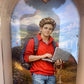 Pormenor do Oratório Carlo Acutis Fiorello com imagem de São Carlo Acutis de camisola vermelha, mochila, computador portátil e terço, sobre uma paisagem campestre.