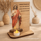 Placa de madeira com imagem do Sagrado Coração de Jesus e vela acesa, com oração religiosa gravada em português.
