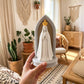 Mão segura um oratório de Nossa Senhora de Fátima com arco cinzento e fundo em madeira, numa sala iluminada com plantas.