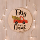 Placa de madeira com o texto "Feliz Natal" em preto, ilustrada com uma carrinha vermelha que transporta uma árvore e presentes. Fundo rústico com luzes decorativas suaves.