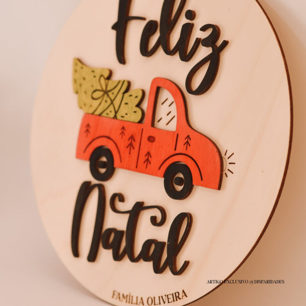 Placa de madeira com detalhe de carrinha vermelha carregando uma árvore de Natal. O texto "Feliz Natal" em preto destaca-se em relevo. Design rústico e personalizado com o nome da família.