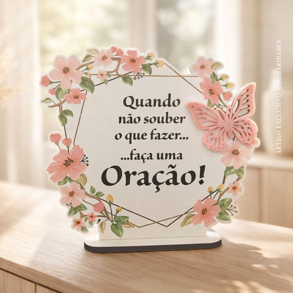 Placa decorativa com a frase “Quando não souber o que fazer… faça uma oração!”, rodeada por flores em tons suaves e uma borboleta rosa em relevo, apoiada sobre uma base de madeira clara, num ambiente luminoso e acolhedor.