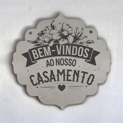 Placa rústica de madeira pendurada com a mensagem "Bem-vindos ao nosso casamento", decorada com flores. Emoldurada por flores naturais em ambiente romântico e acolhedor.